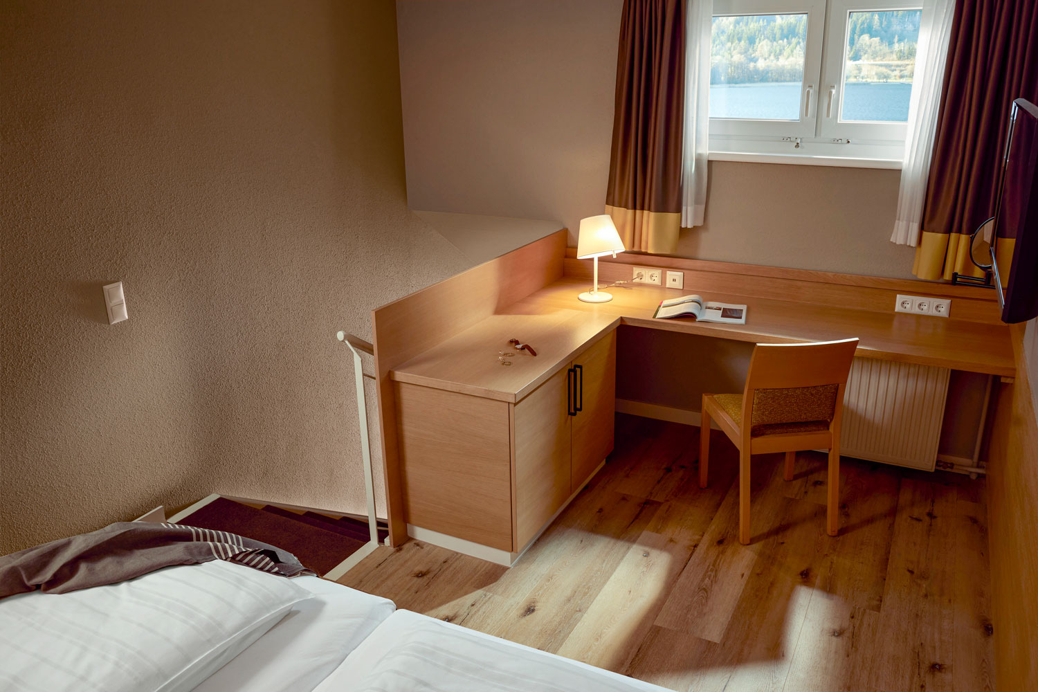 Zimmer mit Holzboden, Schreibtisch am Fenster und einem Bett im Vordergrund